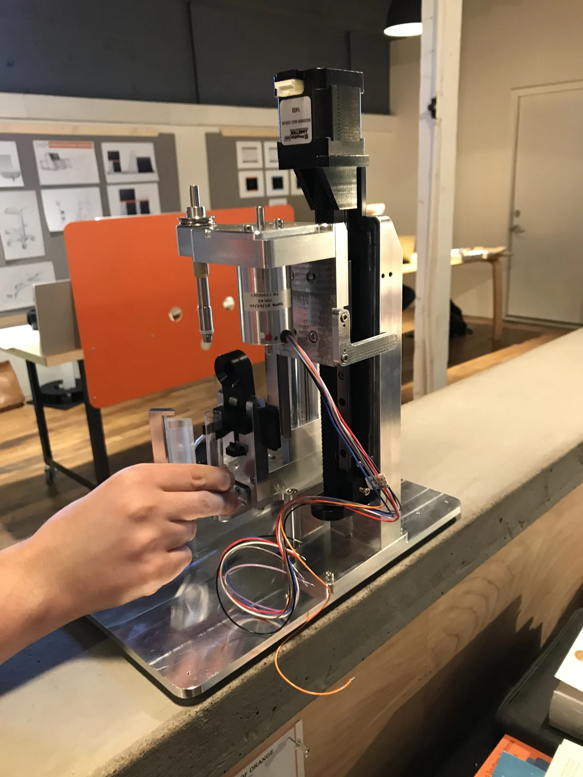 A hand positions a clear component beside a metal prototype with a vertical motor, linear stage, and wiring.