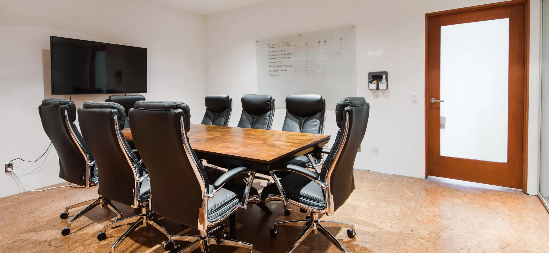 A wooden meeting table, screen, chairs and a project-planning whiteboard.